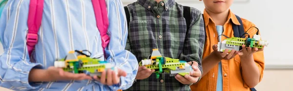 Panoramic crop of multiethnic schoolkids holding robots in classroom — Stock Photo