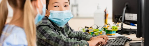 Website header of asian schoolboy in medical mask using computer near classmate in classroom — Stock Photo