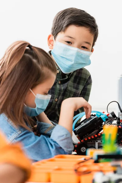 Selektiver Fokus eines asiatischen Schülers in medizinischer Maske, der in der Nähe von Freund Modellierroboter in der Stammschule steht — Stockfoto