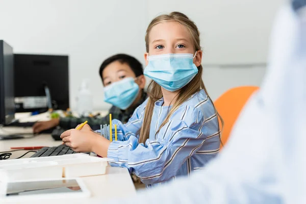 Focus selettivo degli alunni multietnici in maschere mediche seduti vicino ai computer nella scuola staminale — Foto stock
