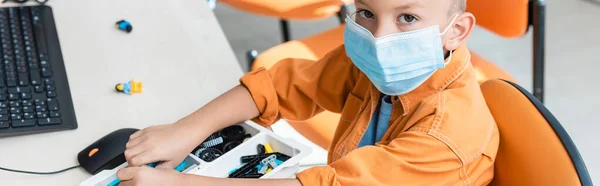 Imagen panorámica del escolar en máscara médica mirando a la cámara cerca de los bloques de construcción y el ordenador - foto de stock