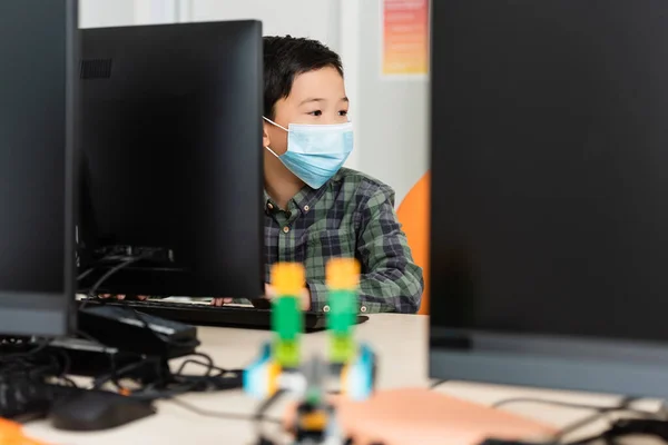 Selective focus of asian schoolboy in medical mask sitting near computers in stem school — Stock Photo