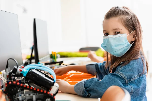 Selektiver Fokus der Schülerin in medizinischer Maske, die neben Robotern und Computern im Klassenzimmer sitzt — Stockfoto
