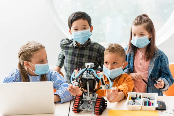 Selective focus of multiethnic classmates in medical masks constructing robot near laptop in classroom — Stock Photo