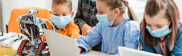 Panoramic shot of schoolkids programming robot near laptop in classroom — Stock Photo