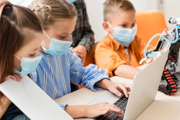 Selective focus of kids in medical masks using laptop and digital tablet near robot in school — Stock Photo