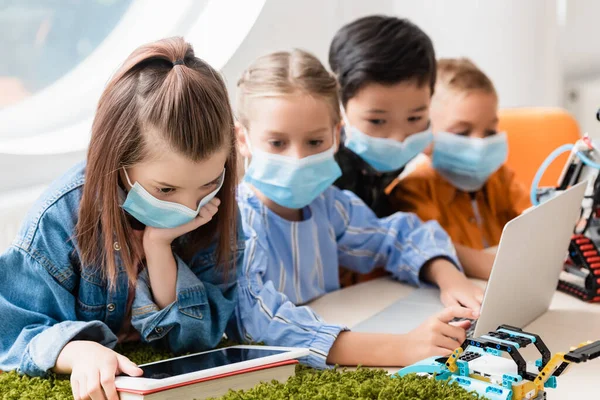 Selective focus of multiethnic schoolkids in medical masks using gadgets while programming robot in stem school — Stock Photo