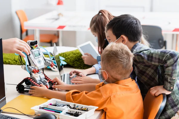 Focus selettivo dell'insegnante che tiene robot vicino agli alunni multietnici in maschere mediche e gadget in classe — Foto stock