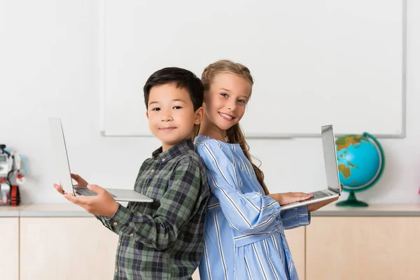 Multicultural pupils holding laptops and looking at camera in classroom — Stock Photo