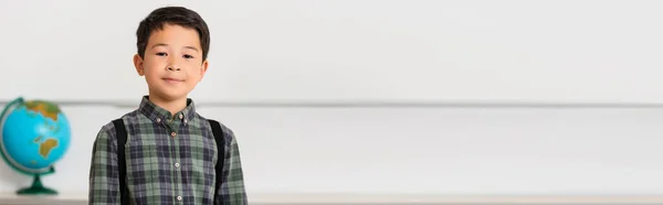 Panoramic shot of asian schoolboy looking at camera in classroom — Stock Photo