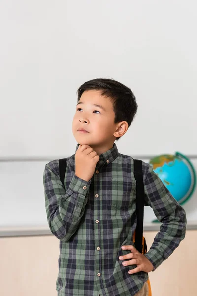 Thoughtful asian schoolboy with hand on hip in classroom — Stock Photo