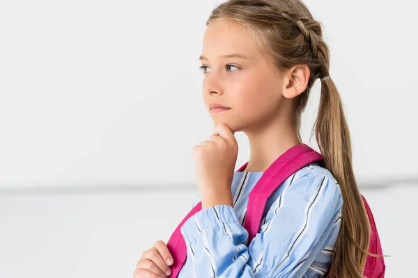 Thoughtful schoolgirl with backpack touching chin in classroom — Stock Photo