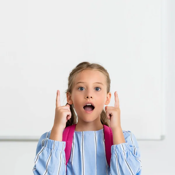 Excited kid with backpack pointing with fingers while looking at camera in school — Stock Photo