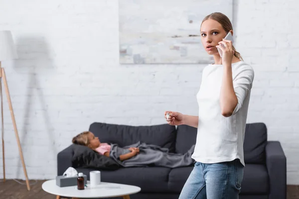 Вибірковий фокус жінки розмовляє на смартфоні і тримає термометр біля хворої дочки на дивані — стокове фото