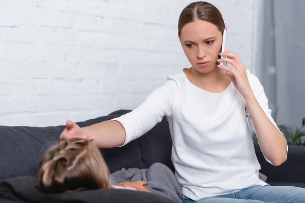 Enfoque selectivo de la mujer joven hablando en el teléfono inteligente y tocando la frente de la hija enferma en el sofá - foto de stock