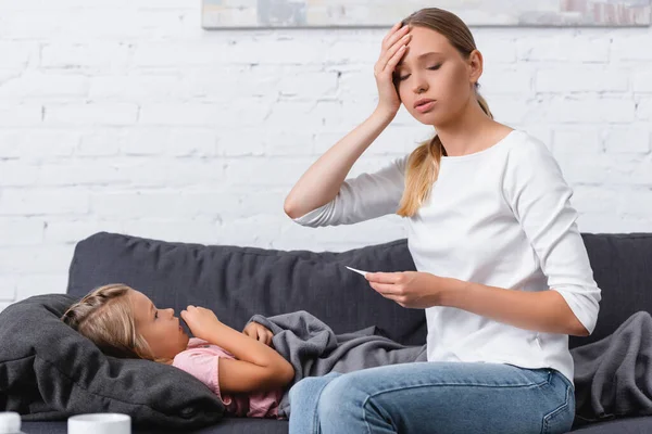 Enfoque selectivo de la mujer enferma sosteniendo el termómetro cerca de la hija en el sofá - foto de stock