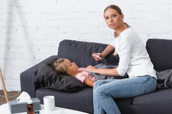 Selective focus of woman with thermometer looking at camera near ill child on sofa — Stock Photo