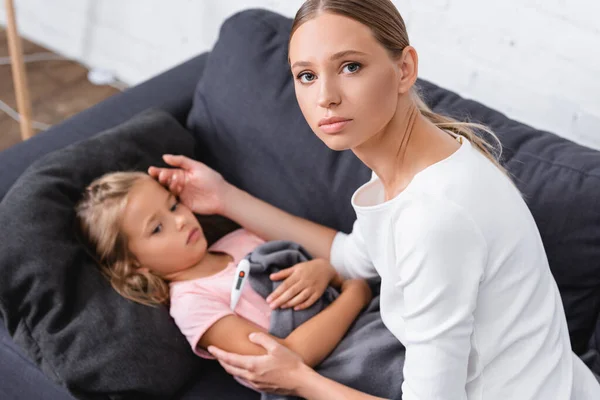Selektiver Fokus der Frau, die in die Kamera schaut, während sie die Stirn ihrer Tochter mit dem Thermometer berührt — Stockfoto