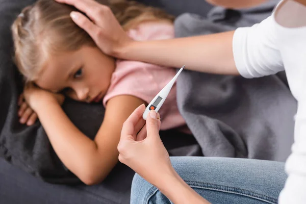 Selektiver Fokus einer Frau mit Thermometer und Streicheln kranker Tochter auf der Couch — Stockfoto