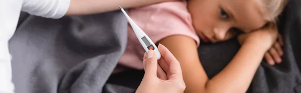 Panoramic shot of woman holding thermometer near ill kid at home — Stock Photo