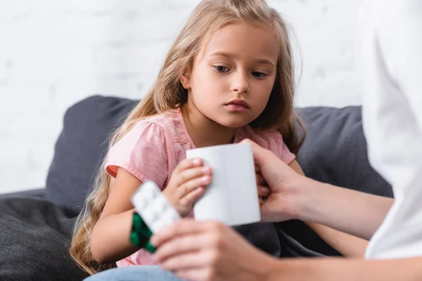 Selektiver Fokus der Frau, die ihrer kranken Tochter eine Tasse schenkt, während sie zu Hause Blasen mit Pillen hält — Stockfoto