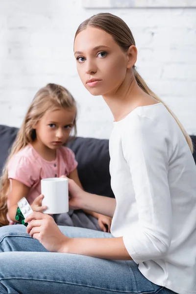 Enfoque selectivo de la mujer joven sosteniendo taza y ampollas con pastillas cerca de la hija enferma en casa - foto de stock