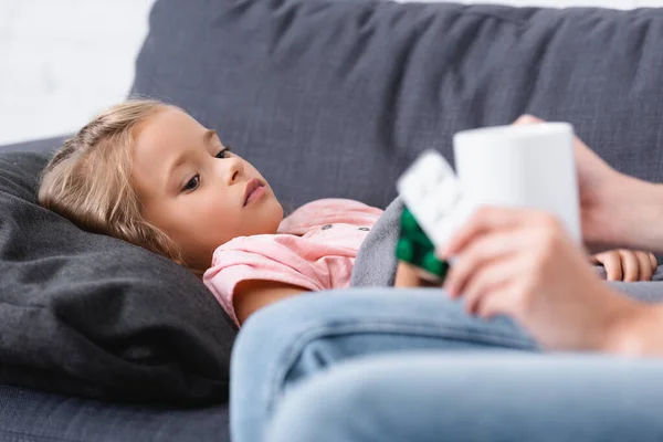 Selektiver Fokus der Frau, die Tasse und Pillen in der Nähe eines kranken Kindes auf der Couch hält — Stockfoto