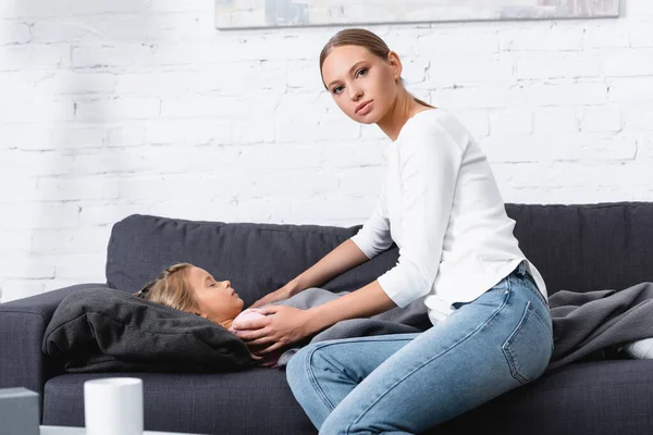 Selective focus of woman looking at camera while touching sick child on couch — Stock Photo