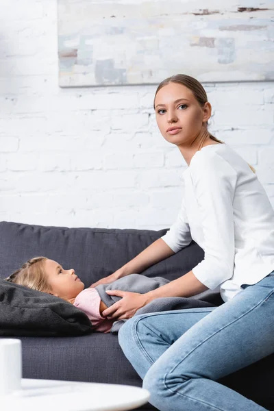 Selektiver Fokus der Frau, die in die Kamera schaut, während sie kranke Tochter auf dem Sofa berührt — Stockfoto