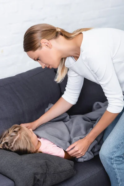 Mutter deckt kranke Tochter zu Hause mit Decke auf Couch ab — Stockfoto