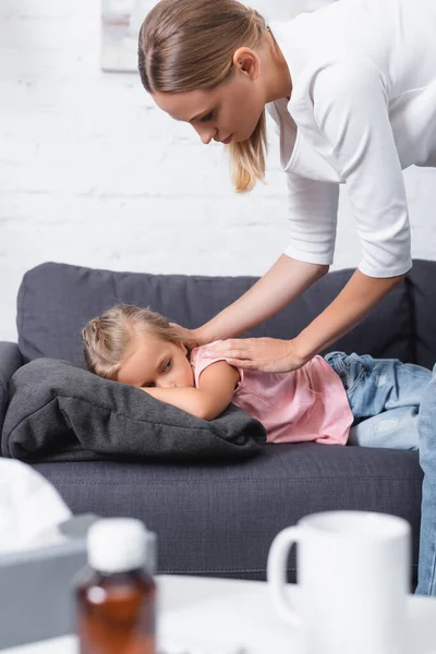 Selektiver Fokus einer jungen Frau, die krankes Kind in der Nähe von Tasse und Servietten auf Couchtisch berührt — Stockfoto