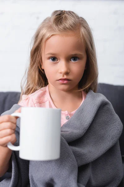 Selektive Fokussierung des Mädchens in einer Decke, die Tasse hält und zu Hause in die Kamera schaut — Stockfoto