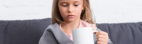 Foto panorámica de niño enfermo en manta mirando taza en casa - foto de stock
