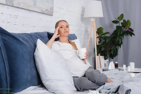 Selektiver Fokus der schwangeren Frau, die den Kopf berührt und Tasse auf dem Bett hält — Stockfoto