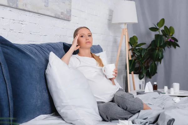 Focus selettivo della donna incinta che tocca la testa e tiene la tazza sul letto — Foto stock