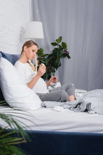 Selektiver Fokus einer Schwangeren, die auf ein Glas mit Pillen blickt und ein Glas Wasser in der Nähe von Servietten auf dem Bett hält — Stockfoto