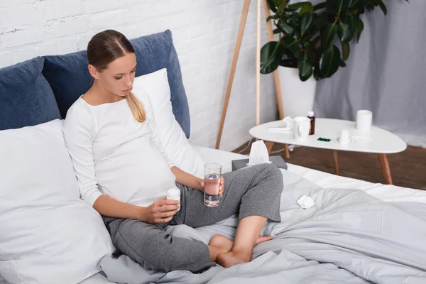 Mujer embarazada enferma sosteniendo un vaso de agua y pastillas mientras está sentada en la cama en casa - foto de stock