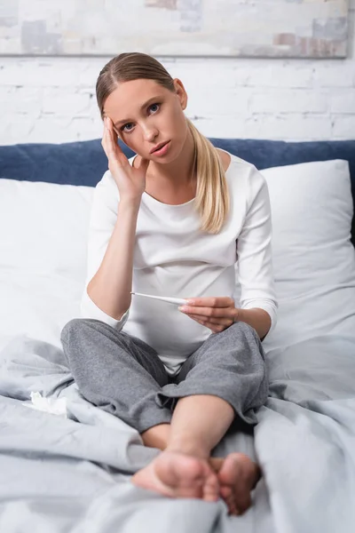 Mise au point sélective de la femme enceinte avec la main près de la tête regardant la caméra tout en tenant le thermomètre sur le lit — Photo de stock