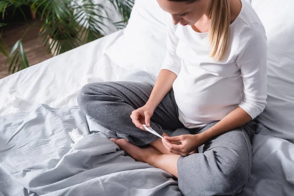 Femme enceinte regardant thermomètre près de serviettes agglomérées sur le lit à la maison — Photo de stock