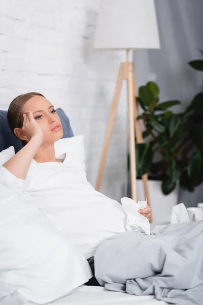 Selective focus of diseased pregnant woman touching head and holding napkin on bed — Stock Photo