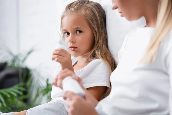 Enfoque selectivo de niña enferma sosteniendo frasco con pastillas cerca de la madre en casa - foto de stock