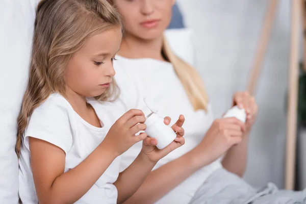 Foco seletivo da menina segurando frasco com pílulas enquanto sentado perto da mãe em casa — Fotografia de Stock