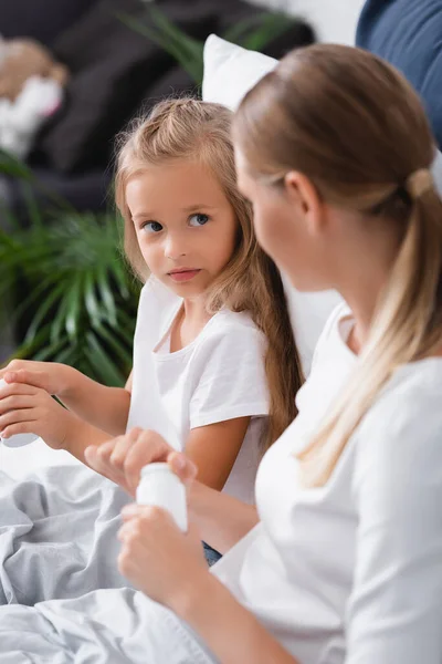 Selective focus of girl looking at mother holding jar of pills on bed at home — Stock Photo