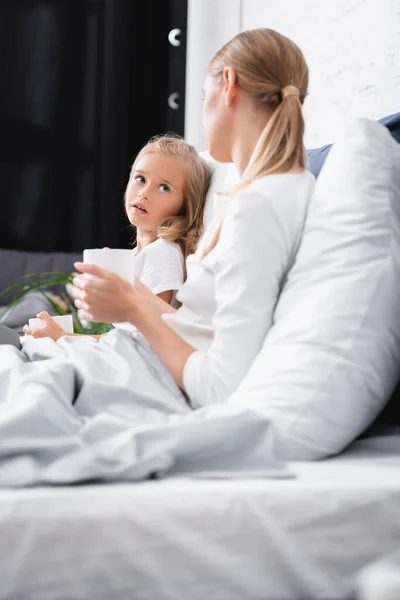 Concentration sélective de la fille tenant tasse et regardant la mère pendant la maladie sur le lit — Photo de stock