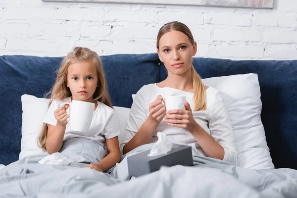 Selective focus of mother and kid looking at camera while holding mugs near box with napkins on bed — Stock Photo