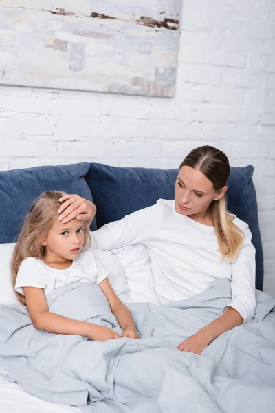 Selective focus of woman touching forehead of sick daughter on bed — Stock Photo