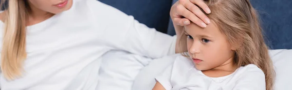 Panoramic shot of young woman touching forehead of diseased daughter on bed — Stock Photo