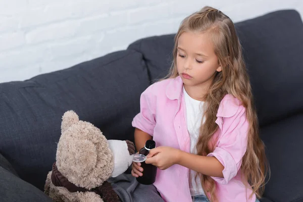 Girl holding spoon and bottle of syrup near soft toy on couch at home — Stock Photo