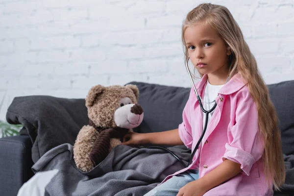 Fille en stéthoscope regardant caméra près de l'ours en peluche sur le canapé — Photo de stock