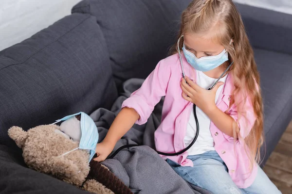 Focus selettivo della ragazza in maschera medica che tiene lo stetoscopio vicino al giocattolo morbido a casa — Foto stock